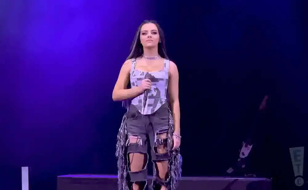 Singer performs on stage in a corset top and ripped shorts with fringe, holding a microphone.