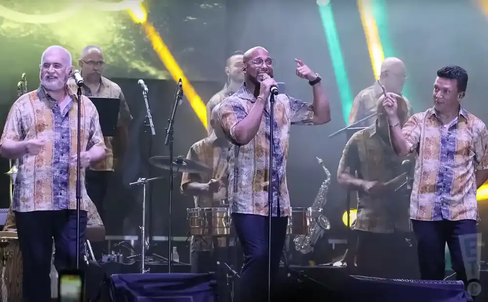 Musicians in patterned shirts perform on stage with instruments and microphones.
