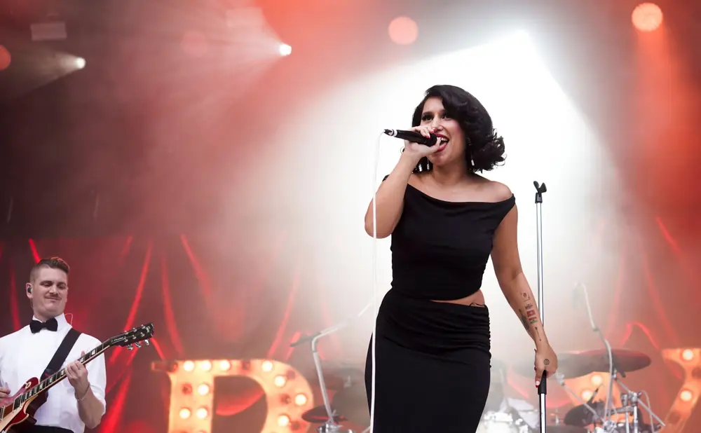 A female singer performs on stage in a black off-the-shoulder dress, with a guitarist in a white shirt playing in the background.