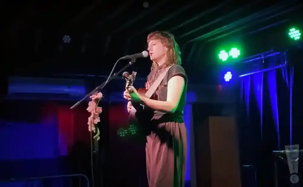 A person with shoulder-length hair plays an acoustic guitar and sings into a microphone on a dimly lit stage.