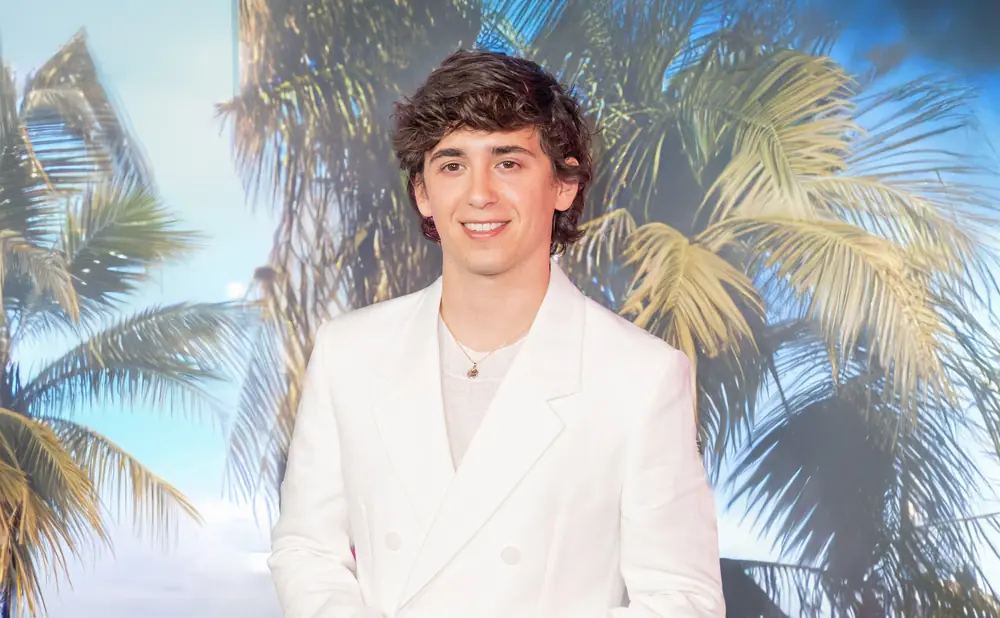 A young man in a white suit smiles at the camera, with a backdrop of palm trees and a sunny sky.