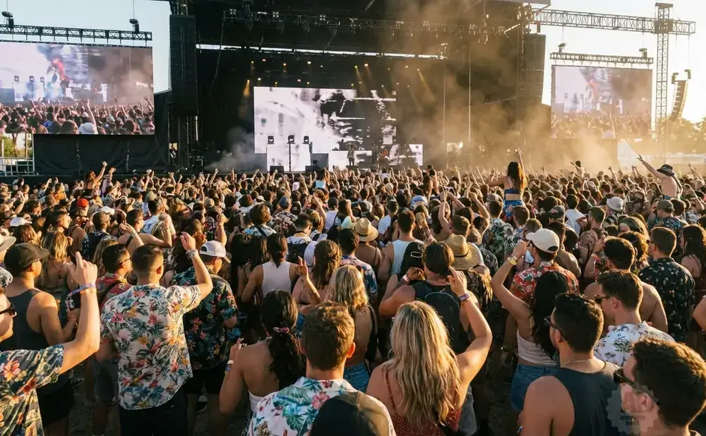 Crowd at an outdoor concert in front of a large stage with screens displaying the performers.