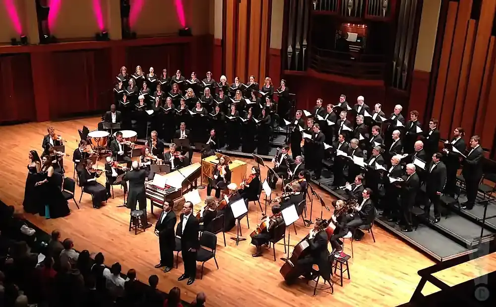 A choir and orchestra perform on a stage, with an organ and audience in the background.