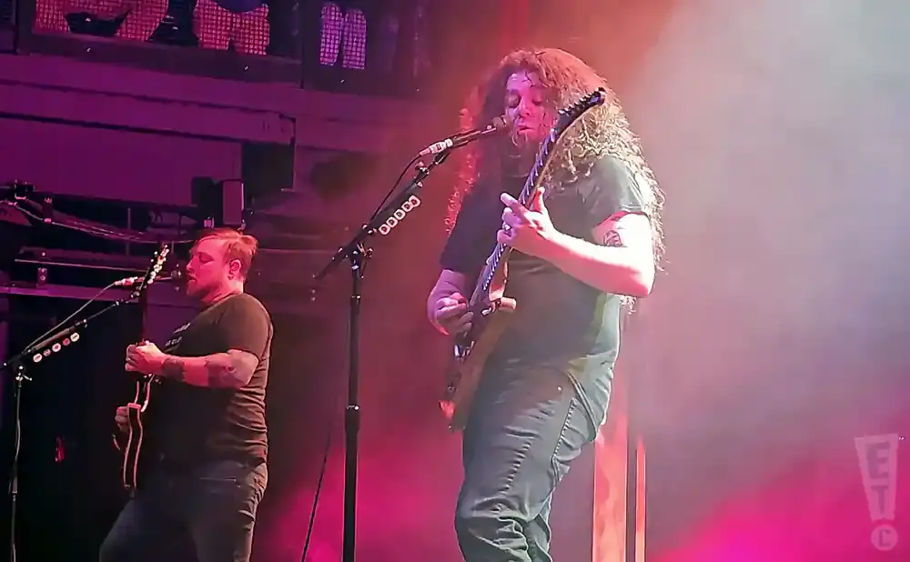 Two men play guitar on a stage with purple and pink lighting.