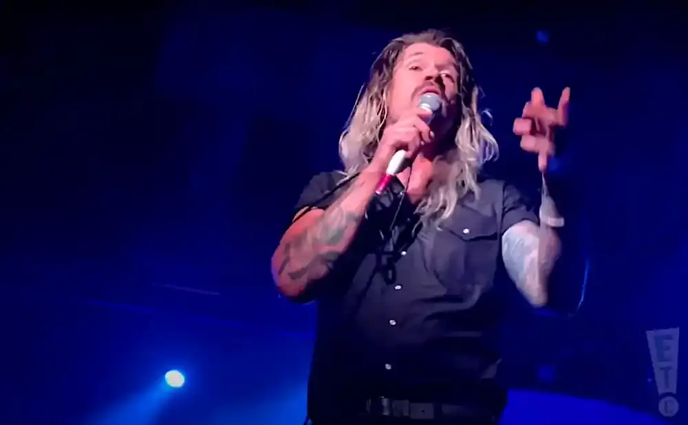 A tattooed man with long blonde hair and a mustache sings into a microphone on a dark, blue-lit stage.
