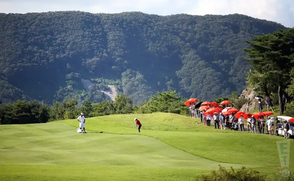 Golfers play a round on a course with a lush mountain backdrop, while spectators hold bright orange umbrellas.