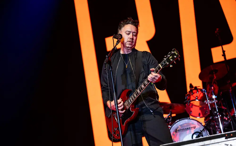 A man in a black jacket plays a red electric guitar on stage with a large orange 