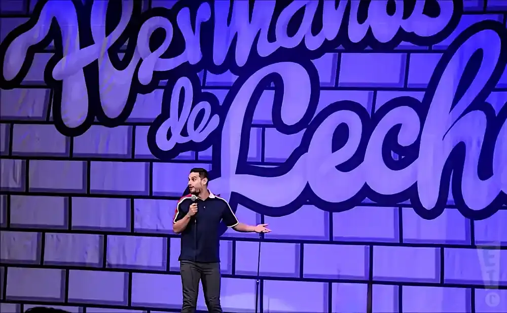 A man tells a joke on stage with a brick-patterned backdrop featuring a large 