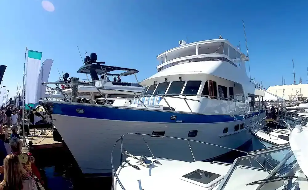 A large white and blue yacht is docked at a sunny outdoor boat show next to other boats and people.