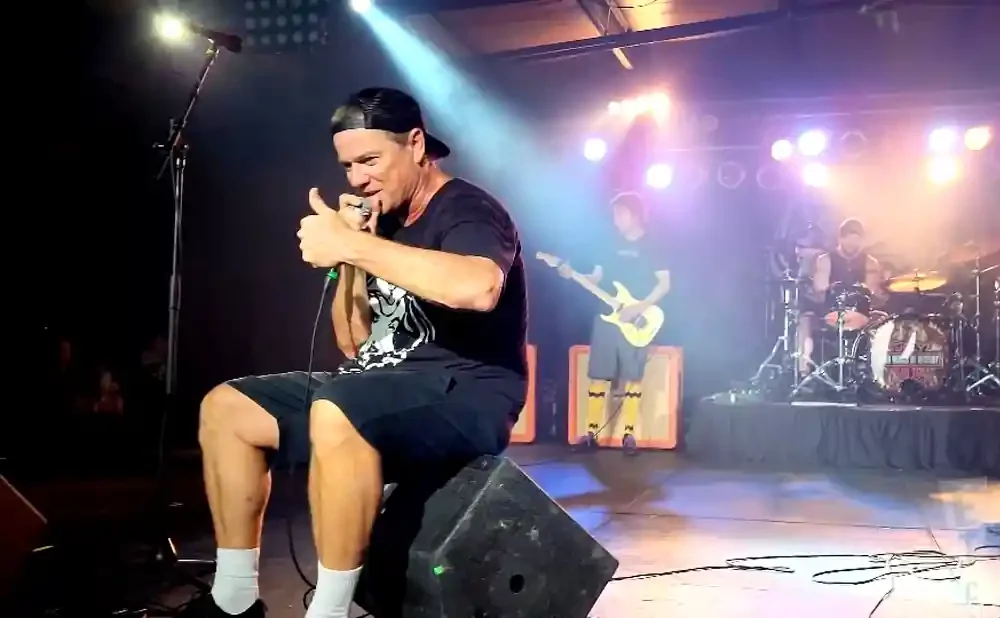 A man in a black cap and t-shirt sits on a speaker, holding a microphone and giving a thumbs-up.