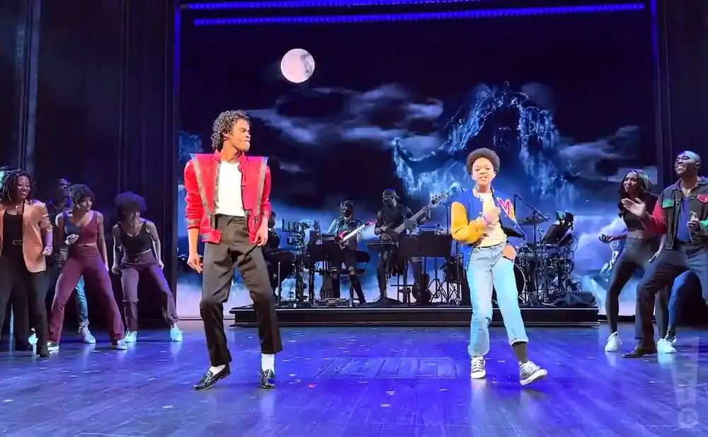 A performer in a red jacket dances on stage with a moon and mountain backdrop.