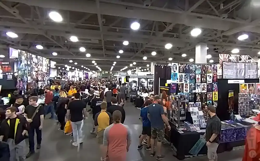 A crowded convention hall with many people browsing tables of artwork and merchandise.