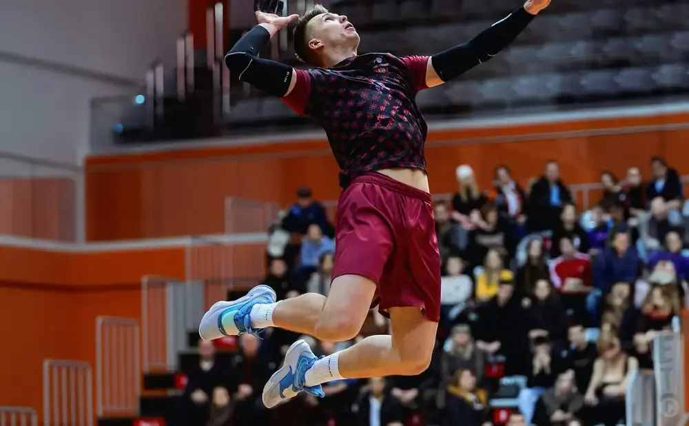 Volleyball player in mid-air during a game, wearing a maroon jersey and shorts, with spectators in the background.