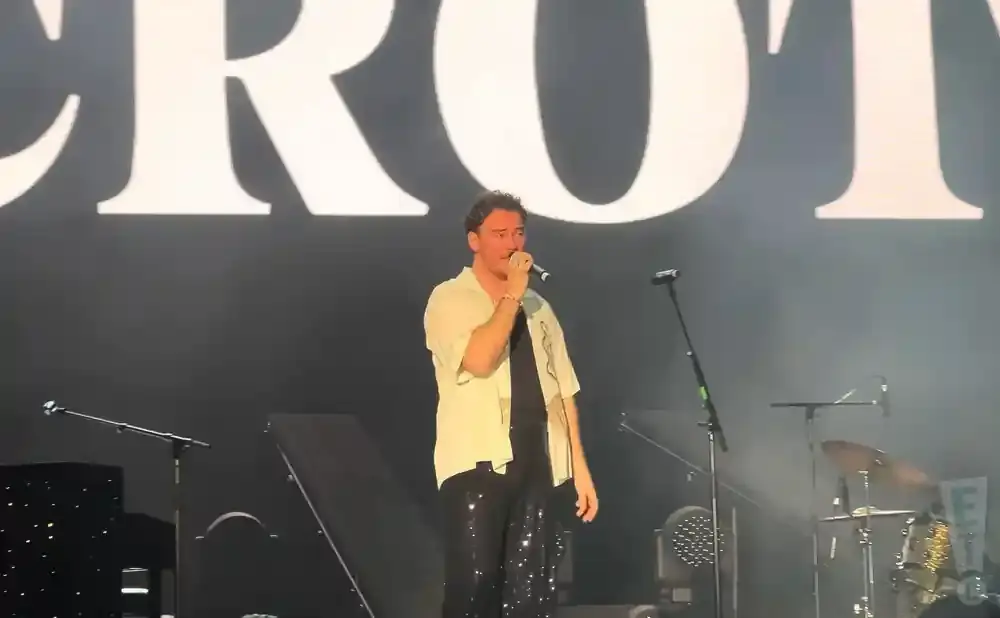A man sings into a microphone on stage, wearing a white shirt and sparkly black pants.