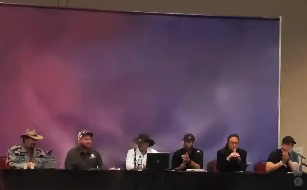 Six men sit at a table in front of a purple and pink backdrop.