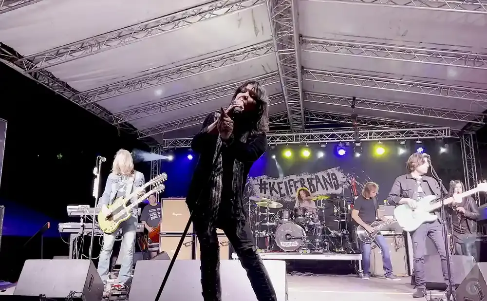 A rock band performs on stage at night under a canopy, with the lead singer in the foreground.