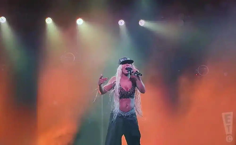 A woman with long blonde hair, wearing a black cap and outfit, sings into a microphone on a stage with orange lighting and smoke effects.