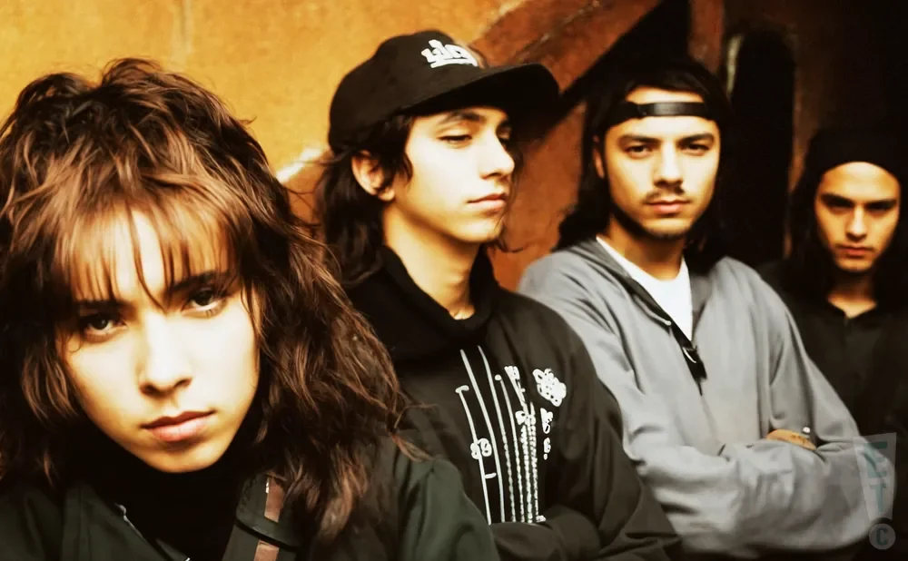 A band of four young people with long hair pose together, with the woman in front looking directly at the camera.
