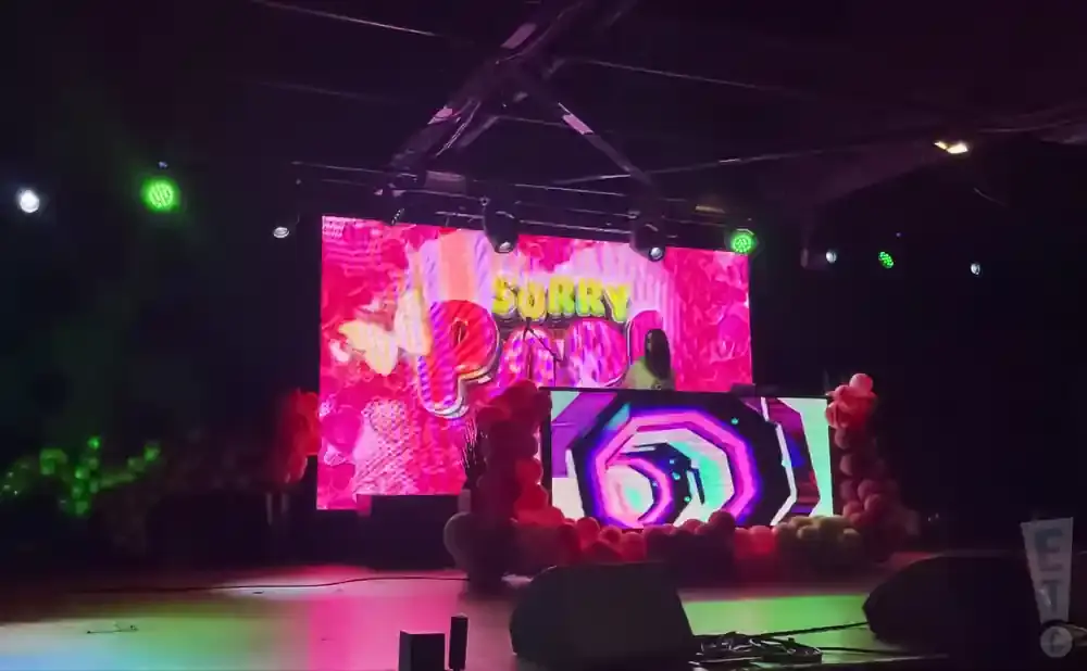 A DJ plays music on a stage with a large pink screen displaying 