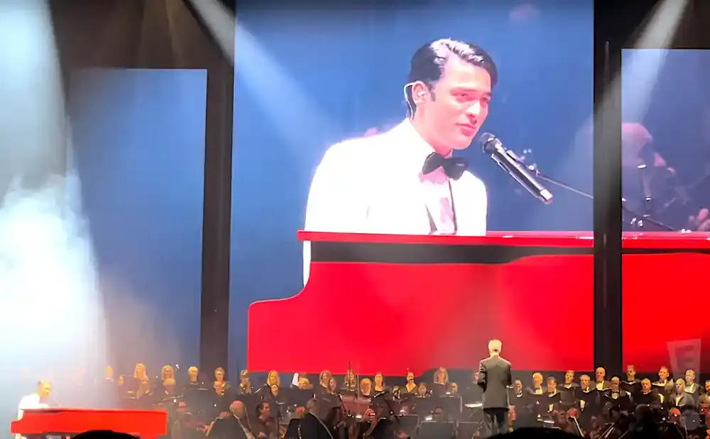 A man in a white suit and bow tie sings at a red piano on a large screen, with a choir and conductor below.