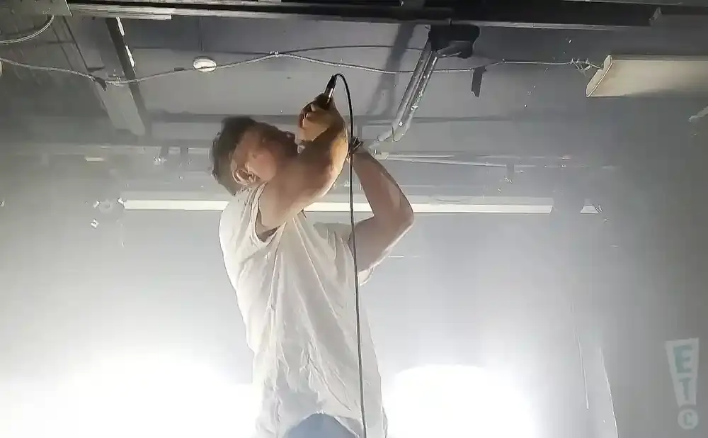 Man in white t-shirt singing into a microphone, on a stage with fog.