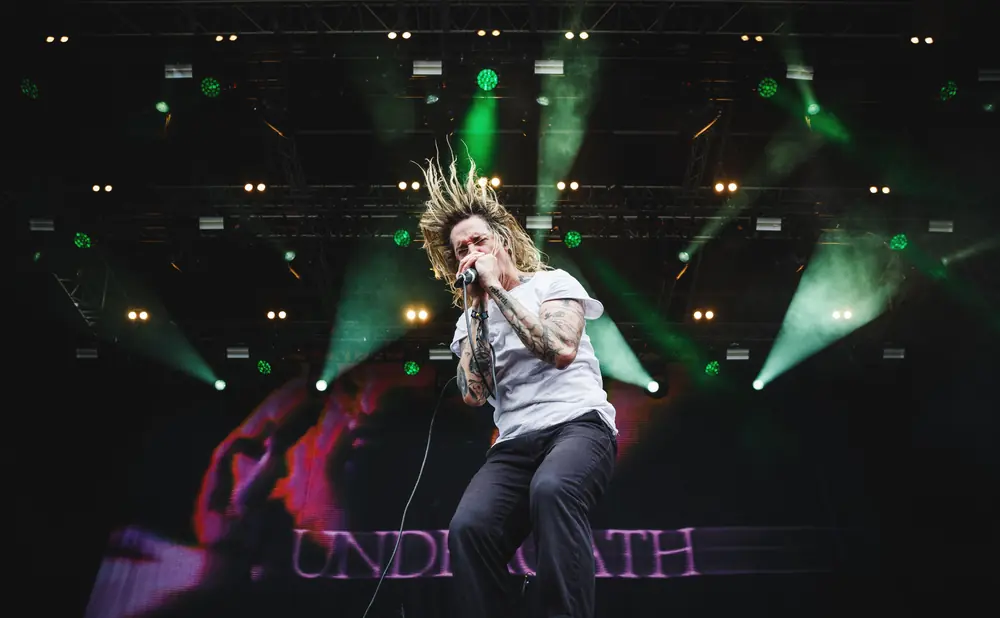 A male singer with wild blonde hair sings into a microphone on a stage with green lights and smoke.