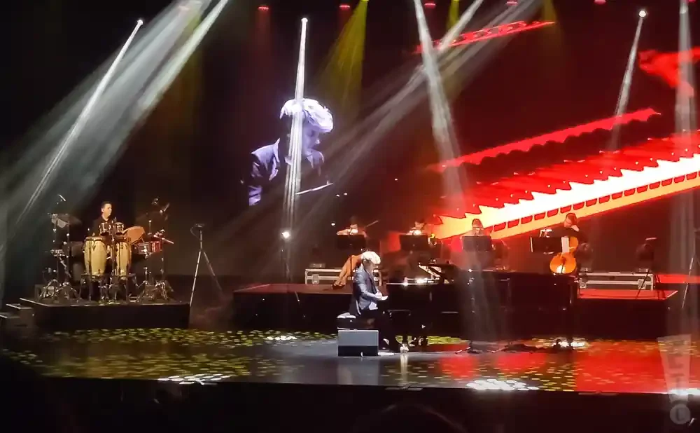 Musicians perform on a stage with bright lights and a large screen showing a performer.