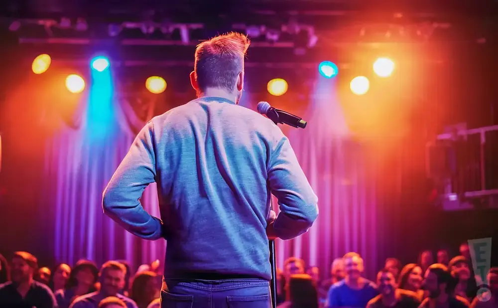 Comedian on stage with a microphone in front of a cheering audience, illuminated by colorful stage lights.