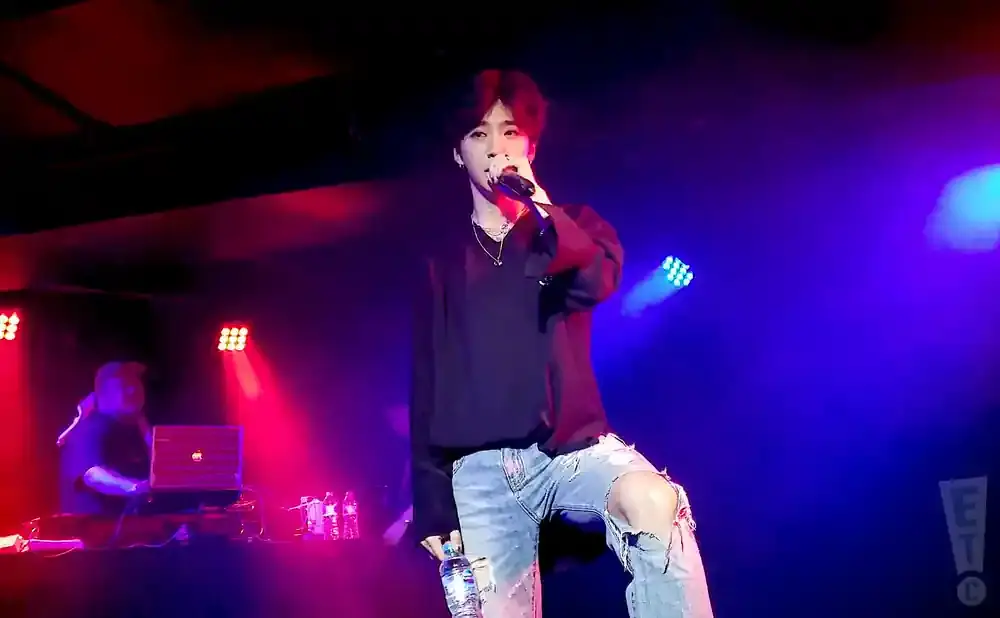 A male singer performs on stage in ripped jeans and a black shirt, holding a water bottle.