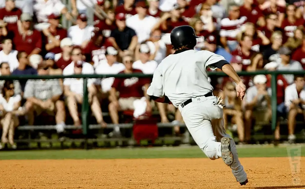 Baseball player in a grey uniform running on a dirt field with spectators in the background.