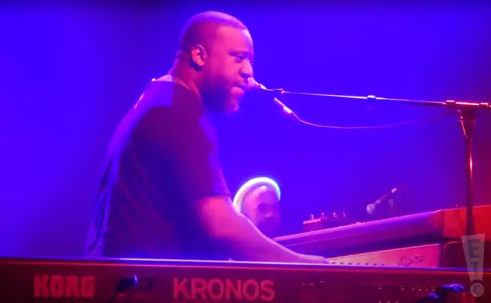 A Black man plays a Korg Kronos keyboard on a stage with blue lighting.