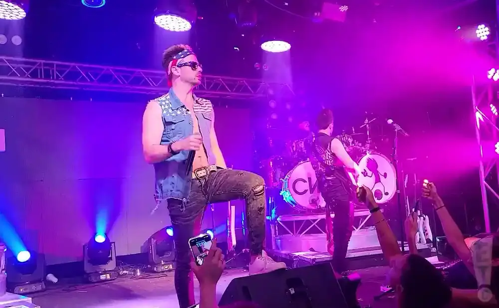 Singer in a denim vest with stars and an American flag bandana performs on stage with bandmates.