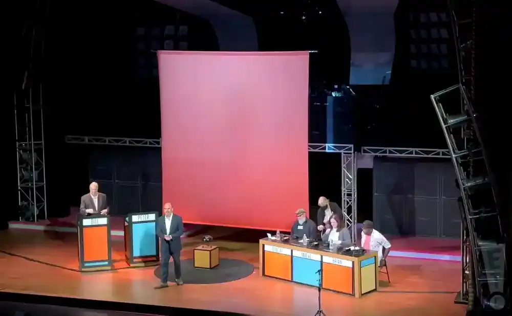 A debate stage with five people on it and two podiums, a large pink screen behind them.