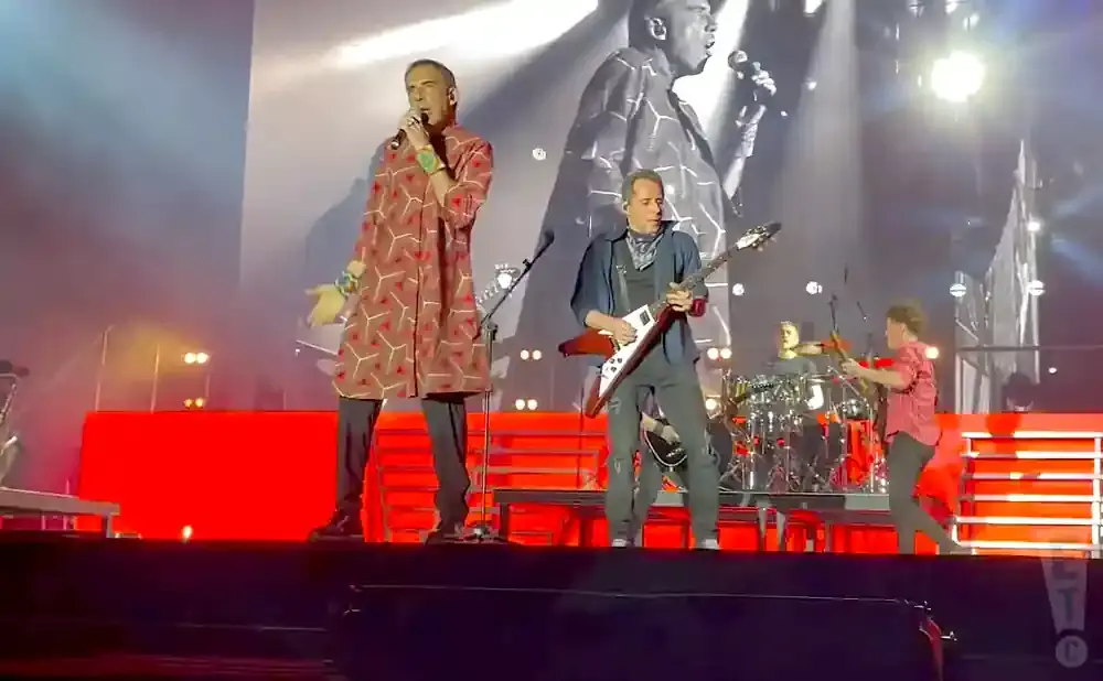 Singer in a patterned tunic and guitarist on stage with a large screen backdrop.