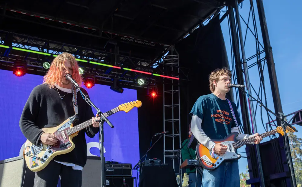 Musicians on a stage play guitars under a blue sky.