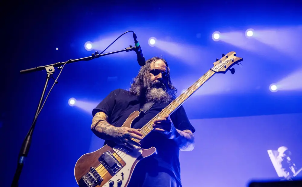 A long-haired, bearded man plays a bass guitar on a stage with blue lighting.