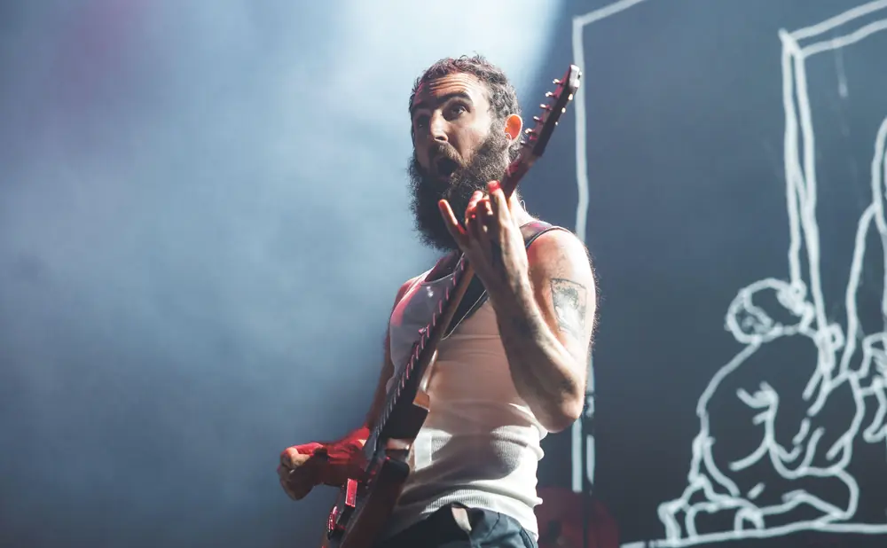 Musician with a beard plays guitar on stage, illuminated by spotlights.