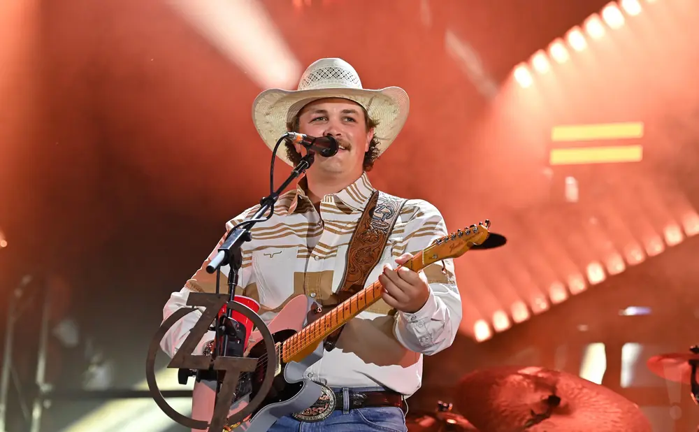 A man in a cowboy hat and striped shirt plays a guitar on stage with warm lighting.