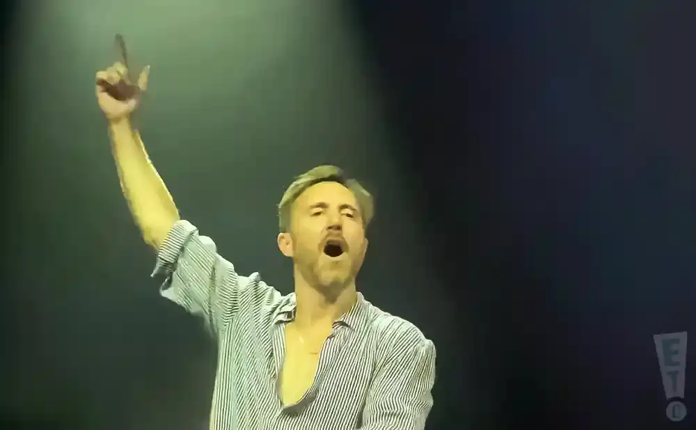 A man with his arm raised and finger pointing upwards sings into a microphone.