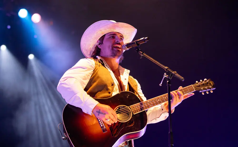 Musician in a cowboy hat sings and plays an acoustic guitar on a dimly lit stage with spotlights.