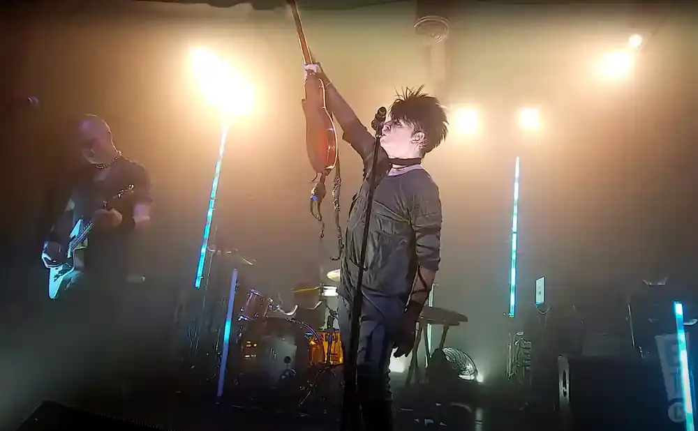 A guitarist holds his guitar aloft on a dimly lit stage with bright lights.
