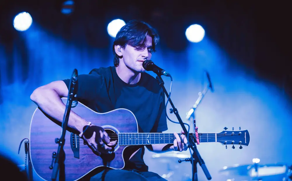A male musician plays an acoustic guitar on a dimly lit stage, bathed in blue light.