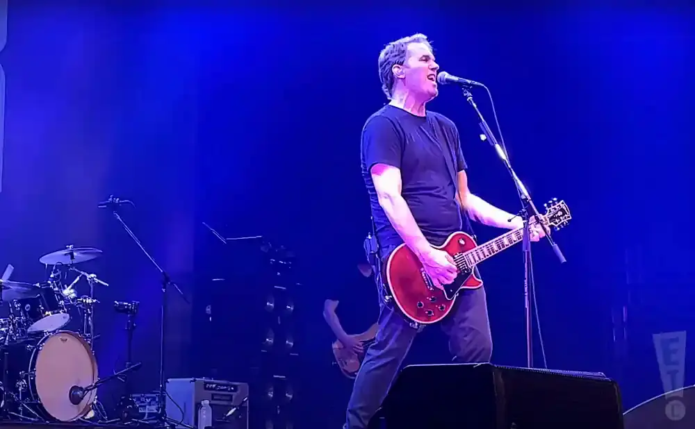 A man in a black t-shirt plays a red electric guitar and sings into a microphone on a stage with blue lighting.