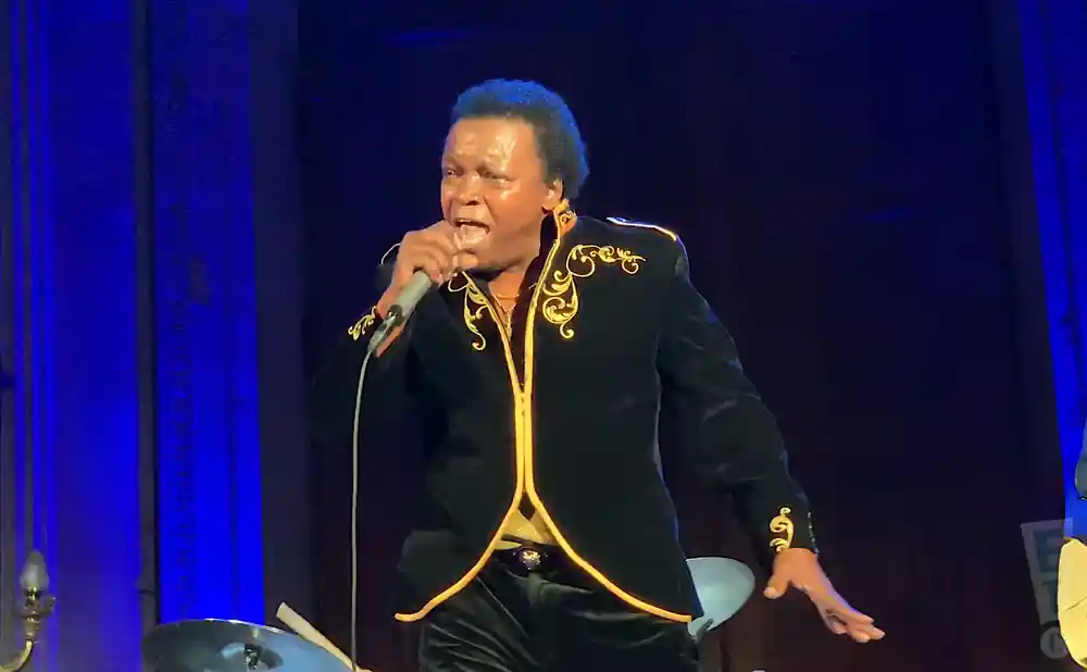 A Black male singer in a black velvet jacket with gold trim sings into a microphone on stage.