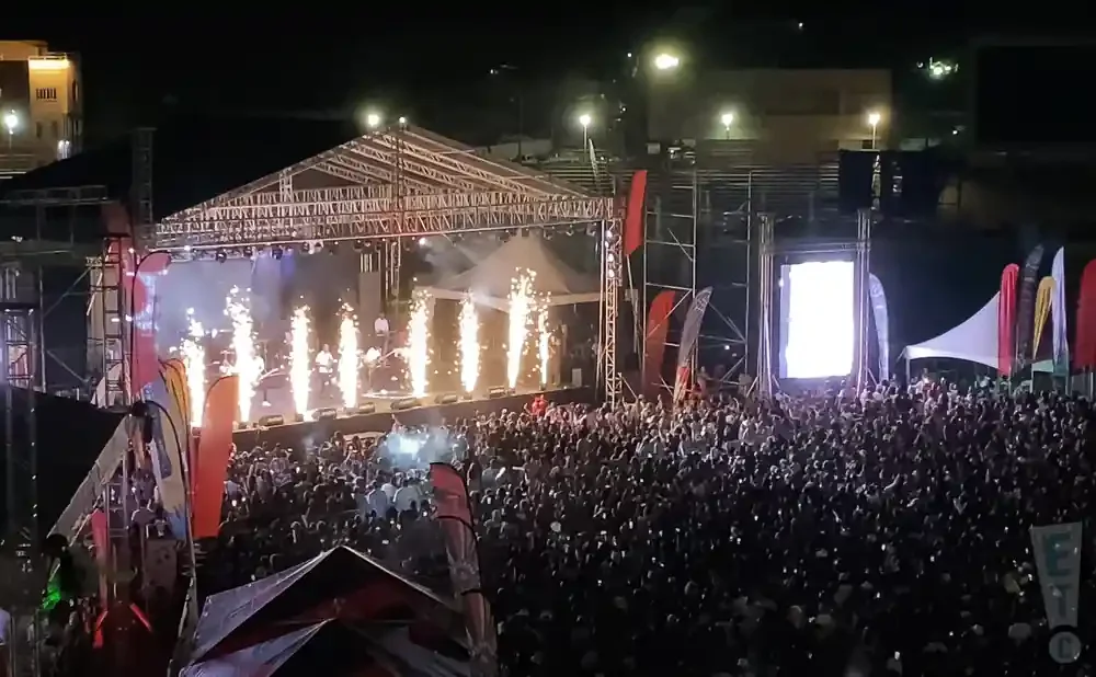 A crowded outdoor concert at night with a stage, pyrotechnics, and a large screen.