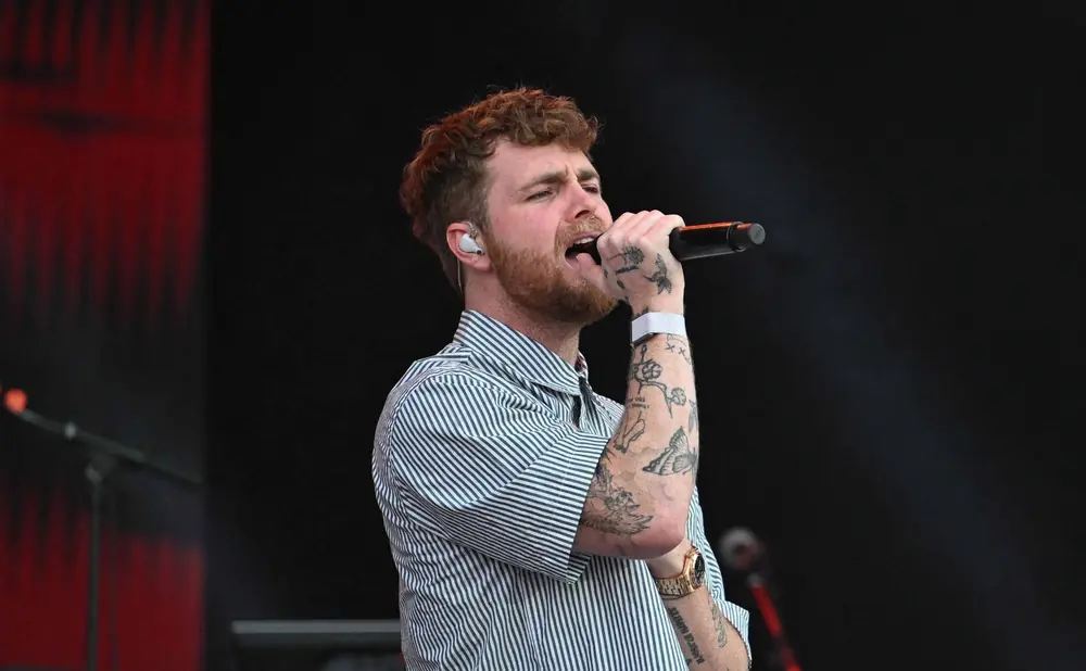 Man with red curly hair sings into a microphone, wearing a striped shirt and has tattoos on his arm.