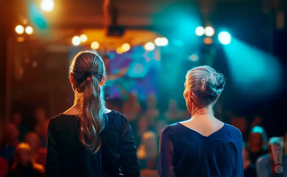 Two women stand facing away from the camera on a dimly lit stage, bathed in colorful spotlights.
