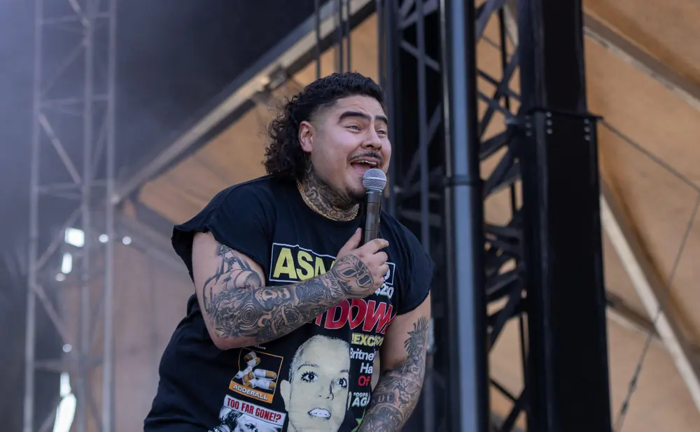A tattooed man with a mullet holds a microphone and smiles onstage in a black t-shirt.
