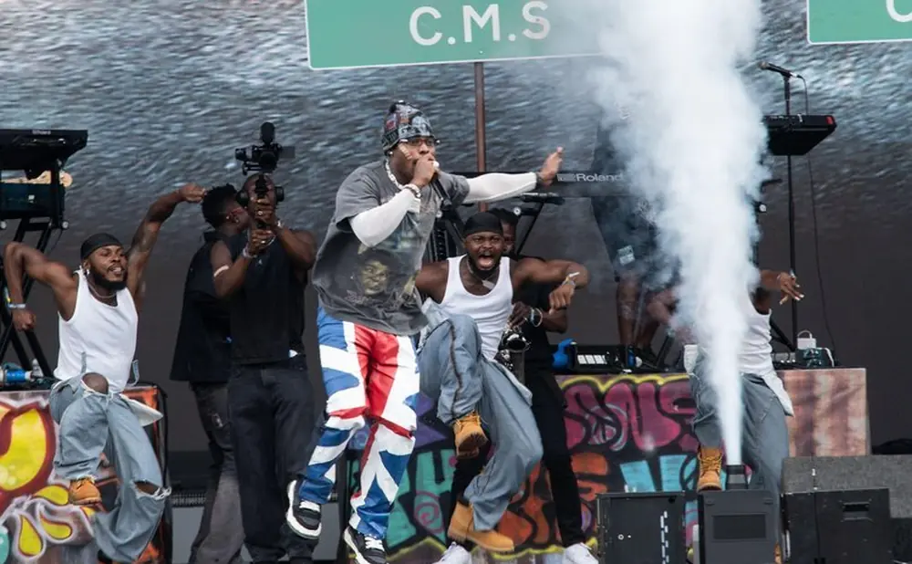A male performer in colorful pants sings on stage with dancers and smoke effects.