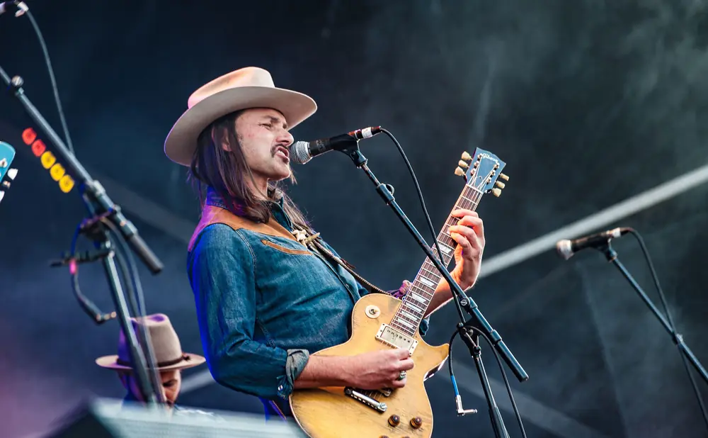A man in a cowboy hat plays a golden electric guitar while singing into a microphone on stage.